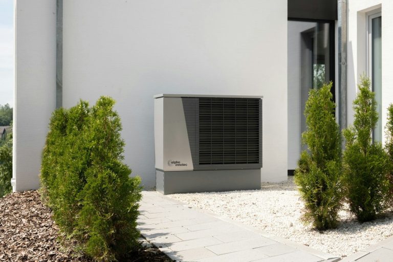 Wärmepumpe, symbolisiert energieeffiziente Sanierung Energieeffiziente Wärmepumpe neben einer modernen Gebäudefassade, umgeben von Pflanzen.