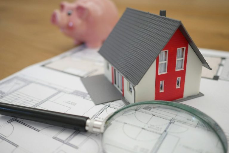 Finanzierungen für Kauf und Neubau von Immobilien Miniaturhausmodell auf Plänen mit einer Lupe und einem Sparschwein im Hintergrund.